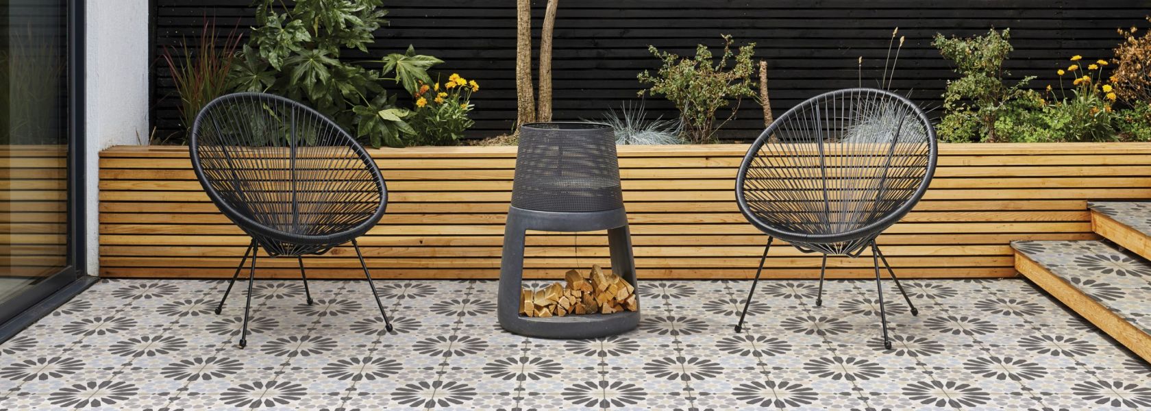 An outdoor seating area with 2 chairs, fire, tiled flooring and wooden plant box and background wall.